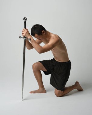 Yakışıklı Asyalı erkek modelin tam boy portresi, spor şortu ve gömleksiz. Kılıç tutan, savaşçı eğitim pozisyonunda diz çöken, beyaz stüdyo arka planında izole edilmiş..