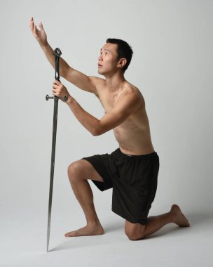 Yakışıklı Asyalı erkek modelin tam boy portresi, spor şortu ve gömleksiz. Kılıç tutan, savaşçı eğitim pozisyonunda diz çöken, beyaz stüdyo arka planında izole edilmiş..