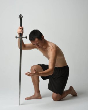 Yakışıklı Asyalı erkek modelin tam boy portresi, spor şortu ve gömleksiz. Kılıç tutan, savaşçı eğitim pozisyonunda diz çöken, beyaz stüdyo arka planında izole edilmiş..