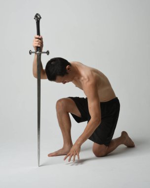 Yakışıklı Asyalı erkek modelin tam boy portresi, spor şortu ve gömleksiz. Kılıç tutan, savaşçı eğitim pozisyonunda diz çöken, beyaz stüdyo arka planında izole edilmiş..