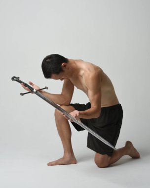 Yakışıklı Asyalı erkek modelin tam boy portresi, spor şortu ve gömleksiz. Kılıç tutan, savaşçı eğitim pozisyonunda diz çöken, beyaz stüdyo arka planında izole edilmiş..