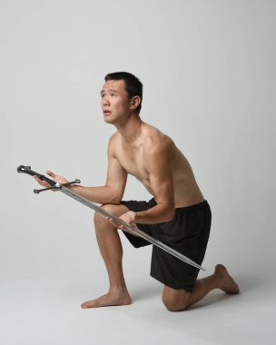 Yakışıklı Asyalı erkek modelin tam boy portresi, spor şortu ve gömleksiz. Kılıç tutan, savaşçı eğitim pozisyonunda diz çöken, beyaz stüdyo arka planında izole edilmiş..