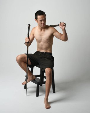 Yakışıklı Asyalı erkek modelin tam boy portresi, spor şortu ve gömleksiz. Kılıç tutan, savaşçı eğitim pozisyonunda oturan, beyaz stüdyo arka planında izole edilmiş.