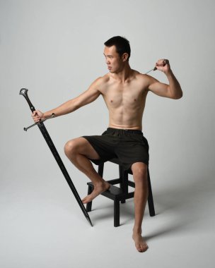 Yakışıklı Asyalı erkek modelin tam boy portresi, spor şortu ve gömleksiz. Kılıç tutan, savaşçı eğitim pozisyonunda oturan, beyaz stüdyo arka planında izole edilmiş.