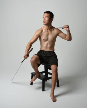Yakışıklı Asyalı erkek modelin tam boy portresi, spor şortu ve gömleksiz. Kılıç tutan, savaşçı eğitim pozisyonunda oturan, beyaz stüdyo arka planında izole edilmiş.