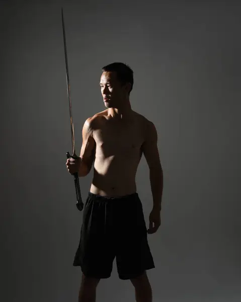 portrait of fit handsome asian male model, wearing gym shorts and shirtless. Holding sword weapons  whilst in warrior training action poses, isolated on moody dark studio background with silhouette