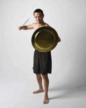 Yakışıklı, tişörtsüz, Asyalı bir erkek modelin tam boy portresi, kılıç ve Spartan kalkanı, savaşçı eğitim pozisyonunda, beyaz stüdyo arka planında izole edilmiş..