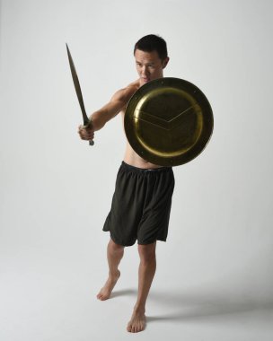 Yakışıklı, tişörtsüz, Asyalı bir erkek modelin tam boy portresi, kılıç ve Spartan kalkanı, savaşçı eğitim pozisyonunda, beyaz stüdyo arka planında izole edilmiş..