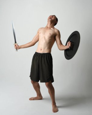 Yakışıklı, tişörtsüz, Asyalı bir erkek modelin tam boy portresi, kılıç ve Spartan kalkanı, savaşçı eğitim pozisyonunda, beyaz stüdyo arka planında izole edilmiş..