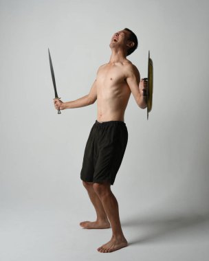 Yakışıklı, tişörtsüz, Asyalı bir erkek modelin tam boy portresi, kılıç ve Spartan kalkanı, savaşçı eğitim pozisyonunda, beyaz stüdyo arka planında izole edilmiş..