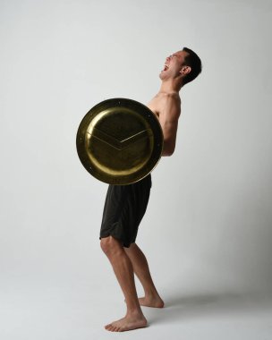 Yakışıklı, tişörtsüz, Asyalı bir erkek modelin tam boy portresi, kılıç ve Spartan kalkanı, savaşçı eğitim pozisyonunda, beyaz stüdyo arka planında izole edilmiş..