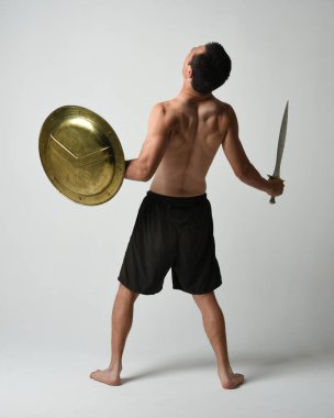 Yakışıklı, tişörtsüz, Asyalı bir erkek modelin tam boy portresi, kılıç ve Spartan kalkanı, savaşçı eğitim pozisyonunda, beyaz stüdyo arka planında izole edilmiş..