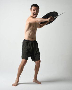 Yakışıklı, tişörtsüz, Asyalı bir erkek modelin tam boy portresi, kılıç ve Spartan kalkanı, savaşçı eğitim pozisyonunda, beyaz stüdyo arka planında izole edilmiş..