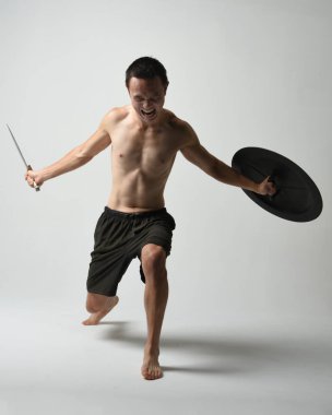 Yakışıklı, tişörtsüz, Asyalı bir erkek modelin tam boy portresi, kılıç ve Spartan kalkanı, savaşçı eğitim pozisyonunda, beyaz stüdyo arka planında izole edilmiş..