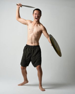 Yakışıklı, tişörtsüz, Asyalı bir erkek modelin tam boy portresi, kılıç ve Spartan kalkanı, savaşçı eğitim pozisyonunda, beyaz stüdyo arka planında izole edilmiş..