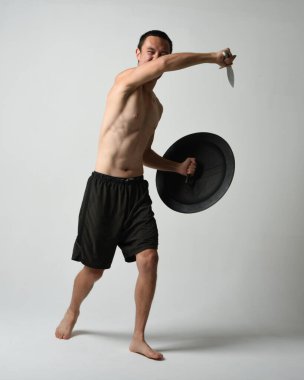 Yakışıklı, tişörtsüz, Asyalı bir erkek modelin tam boy portresi, kılıç ve Spartan kalkanı, savaşçı eğitim pozisyonunda, beyaz stüdyo arka planında izole edilmiş..