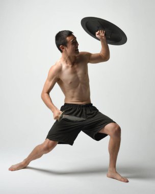 Yakışıklı, tişörtsüz, Asyalı bir erkek modelin tam boy portresi, kılıç ve Spartan kalkanı, savaşçı eğitim pozisyonunda, beyaz stüdyo arka planında izole edilmiş..