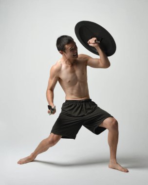 Yakışıklı, tişörtsüz, Asyalı bir erkek modelin tam boy portresi, kılıç ve Spartan kalkanı, savaşçı eğitim pozisyonunda, beyaz stüdyo arka planında izole edilmiş..