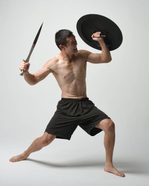 Yakışıklı, tişörtsüz, Asyalı bir erkek modelin tam boy portresi, kılıç ve Spartan kalkanı, savaşçı eğitim pozisyonunda, beyaz stüdyo arka planında izole edilmiş..