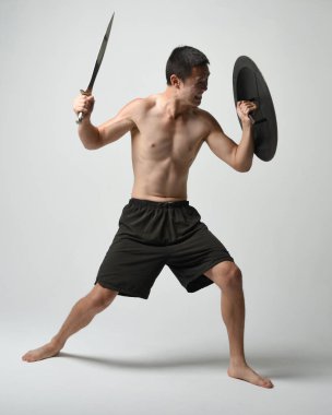 Yakışıklı, tişörtsüz, Asyalı bir erkek modelin tam boy portresi, kılıç ve Spartan kalkanı, savaşçı eğitim pozisyonunda, beyaz stüdyo arka planında izole edilmiş..