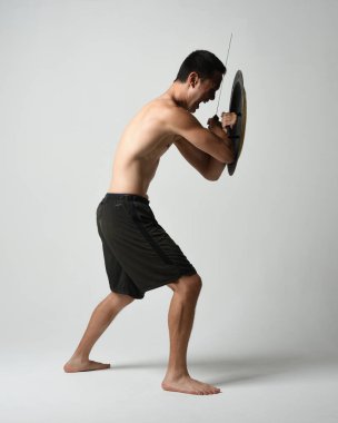 Yakışıklı, tişörtsüz, Asyalı bir erkek modelin tam boy portresi, kılıç ve Spartan kalkanı, savaşçı eğitim pozisyonunda, beyaz stüdyo arka planında izole edilmiş..