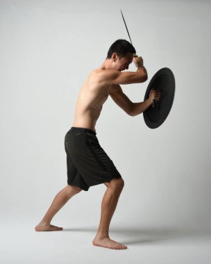 Yakışıklı, tişörtsüz, Asyalı bir erkek modelin tam boy portresi, kılıç ve Spartan kalkanı, savaşçı eğitim pozisyonunda, beyaz stüdyo arka planında izole edilmiş..