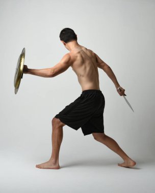 Yakışıklı, tişörtsüz, Asyalı bir erkek modelin tam boy portresi, kılıç ve Spartan kalkanı, savaşçı eğitim pozisyonunda, beyaz stüdyo arka planında izole edilmiş..