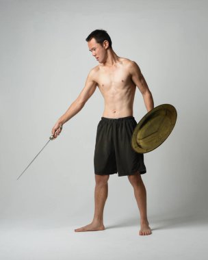 Yakışıklı, tişörtsüz, Asyalı bir erkek modelin tam boy portresi, kılıç ve Spartan kalkanı, savaşçı eğitim pozisyonunda, beyaz stüdyo arka planında izole edilmiş..