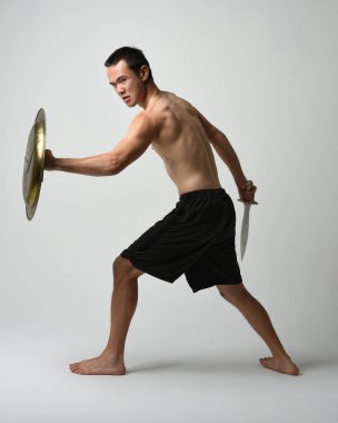 Yakışıklı, tişörtsüz, Asyalı bir erkek modelin tam boy portresi, kılıç ve Spartan kalkanı, savaşçı eğitim pozisyonunda, beyaz stüdyo arka planında izole edilmiş..