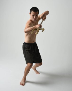 Yakışıklı, tişörtsüz Asyalı bir erkek modelin tam boy portresi, altın mızrak taşıyor, savaşçı eğitim pozisyonunda duruyor, beyaz stüdyo arka planında izole edilmiş..