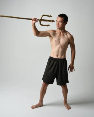 Yakışıklı, tişörtsüz Asyalı bir erkek modelin tam boy portresi, altın mızrak taşıyor, savaşçı eğitim pozisyonunda duruyor, beyaz stüdyo arka planında izole edilmiş..