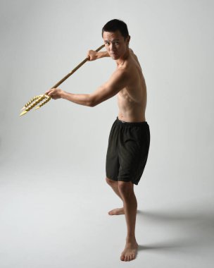 Yakışıklı, tişörtsüz Asyalı bir erkek modelin tam boy portresi, altın mızrak taşıyor, savaşçı eğitim pozisyonunda duruyor, beyaz stüdyo arka planında izole edilmiş..