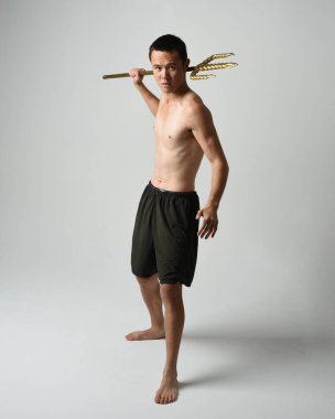 Yakışıklı, tişörtsüz Asyalı bir erkek modelin tam boy portresi, altın mızrak taşıyor, savaşçı eğitim pozisyonunda duruyor, beyaz stüdyo arka planında izole edilmiş..