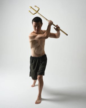 Yakışıklı, tişörtsüz Asyalı bir erkek modelin tam boy portresi, altın mızrak taşıyor, savaşçı eğitim pozisyonunda duruyor, beyaz stüdyo arka planında izole edilmiş..