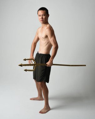 Yakışıklı, tişörtsüz Asyalı bir erkek modelin tam boy portresi, altın mızrak taşıyor, savaşçı eğitim pozisyonunda duruyor, beyaz stüdyo arka planında izole edilmiş..