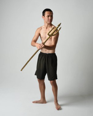 Yakışıklı, tişörtsüz Asyalı bir erkek modelin tam boy portresi, altın mızrak taşıyor, savaşçı eğitim pozisyonunda duruyor, beyaz stüdyo arka planında izole edilmiş..