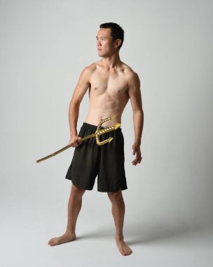 Yakışıklı, tişörtsüz Asyalı bir erkek modelin tam boy portresi, altın mızrak taşıyor, savaşçı eğitim pozisyonunda duruyor, beyaz stüdyo arka planında izole edilmiş..