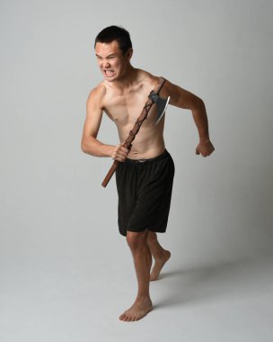 Yakışıklı, tişörtsüz Asyalı erkek modelin tam boy portresi, elinde viking baltası olan, savaşçı eğitim pozisyonunda duran, beyaz stüdyo arka planında izole edilmiş.