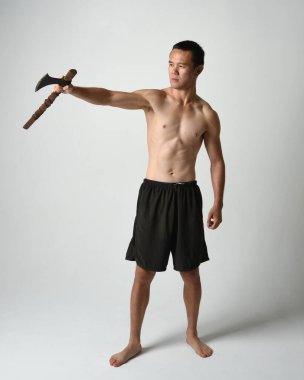 Yakışıklı, tişörtsüz Asyalı erkek modelin tam boy portresi, elinde viking baltası olan, savaşçı eğitim pozisyonunda duran, beyaz stüdyo arka planında izole edilmiş.