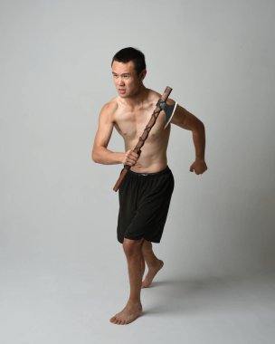 Yakışıklı, tişörtsüz Asyalı erkek modelin tam boy portresi, elinde viking baltası olan, savaşçı eğitim pozisyonunda duran, beyaz stüdyo arka planında izole edilmiş.