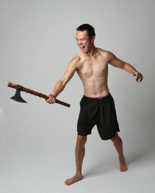 Yakışıklı, tişörtsüz Asyalı erkek modelin tam boy portresi, elinde viking baltası olan, savaşçı eğitim pozisyonunda duran, beyaz stüdyo arka planında izole edilmiş.