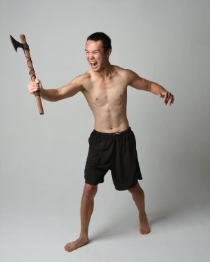 Yakışıklı, tişörtsüz Asyalı erkek modelin tam boy portresi, elinde viking baltası olan, savaşçı eğitim pozisyonunda duran, beyaz stüdyo arka planında izole edilmiş.