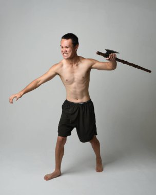 Yakışıklı, tişörtsüz Asyalı erkek modelin tam boy portresi, elinde viking baltası olan, savaşçı eğitim pozisyonunda duran, beyaz stüdyo arka planında izole edilmiş.
