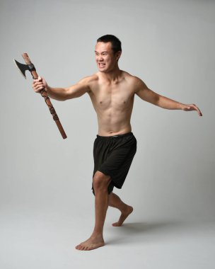 Yakışıklı, tişörtsüz Asyalı erkek modelin tam boy portresi, elinde viking baltası olan, savaşçı eğitim pozisyonunda duran, beyaz stüdyo arka planında izole edilmiş.