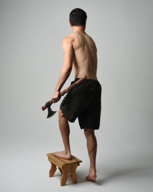 Yakışıklı, tişörtsüz Asyalı erkek modelin tam boy portresi, elinde viking baltası olan, savaşçı eğitim pozisyonunda duran, beyaz stüdyo arka planında izole edilmiş.