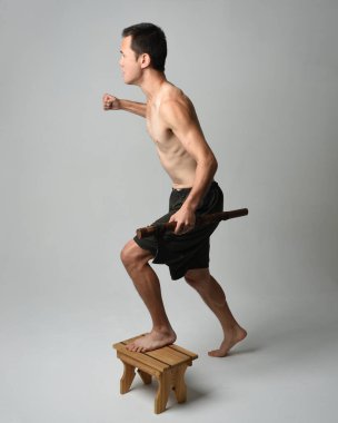 Yakışıklı, tişörtsüz Asyalı erkek modelin tam boy portresi, elinde viking baltası olan, savaşçı eğitim pozisyonunda duran, beyaz stüdyo arka planında izole edilmiş.