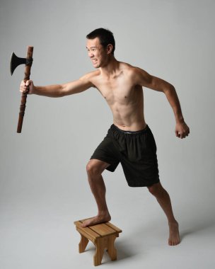 Yakışıklı, tişörtsüz Asyalı erkek modelin tam boy portresi, elinde viking baltası olan, savaşçı eğitim pozisyonunda duran, beyaz stüdyo arka planında izole edilmiş.