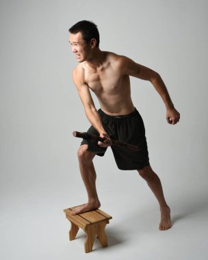 Yakışıklı, tişörtsüz Asyalı erkek modelin tam boy portresi, elinde viking baltası olan, savaşçı eğitim pozisyonunda duran, beyaz stüdyo arka planında izole edilmiş.