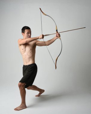 Asyalı erkek modelin tam boy portresi, av yayı ve okçuluk silahı taşıyor, savaşçı eğitim pozisyonunda duruyor, beyaz stüdyo arka planında izole edilmiş..
