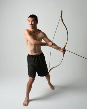 Asyalı erkek modelin tam boy portresi, av yayı ve okçuluk silahı taşıyor, savaşçı eğitim pozisyonunda duruyor, beyaz stüdyo arka planında izole edilmiş..